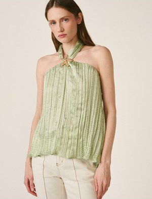 Mujer con un top halter plisado verde claro con un broche en forma de estrella de mar, combinando con pantalones blancos, posando frente a un fondo liso.