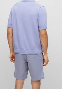 Man wearing light purple short-sleeve shirt and matching shorts, standing facing away against a plain light background.