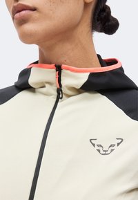 Κοντινό πλάνο ενός ατόμου που φοράει μπεζ και μαύρο μπουφάν με φερμουάρ, με ένα μικρό λογότυπο ζώου στο στήθος και κοραλλί περίγραμμα γύρω από τον γιακά.