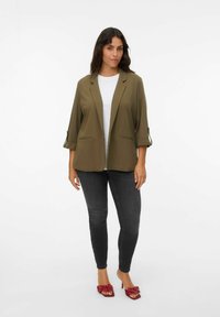 Olivengrüner Blazer mit Dreiviertelärmeln, Reverskragen und Taschen an der Vorderseite, getragen über einem weißen Hemd und kombiniert mit dunklen Jeans.