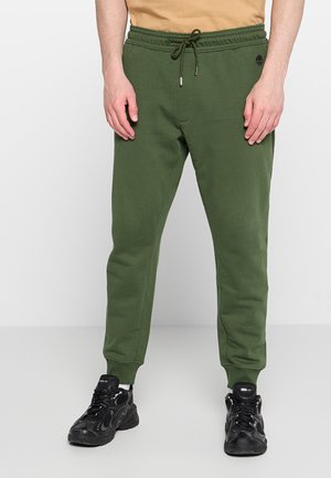 Uomo che indossa pantaloni della tuta verdi con coulisse e scarpe da ginnastica nere, in piedi su sfondo bianco, mani rilassate ai lati.