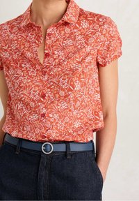 Camicia a maniche corte, con bottoni, di colore arancione con motivo floreale bianco. Abbinata a jeans scuri e una cintura blu con fibbia rotonda.