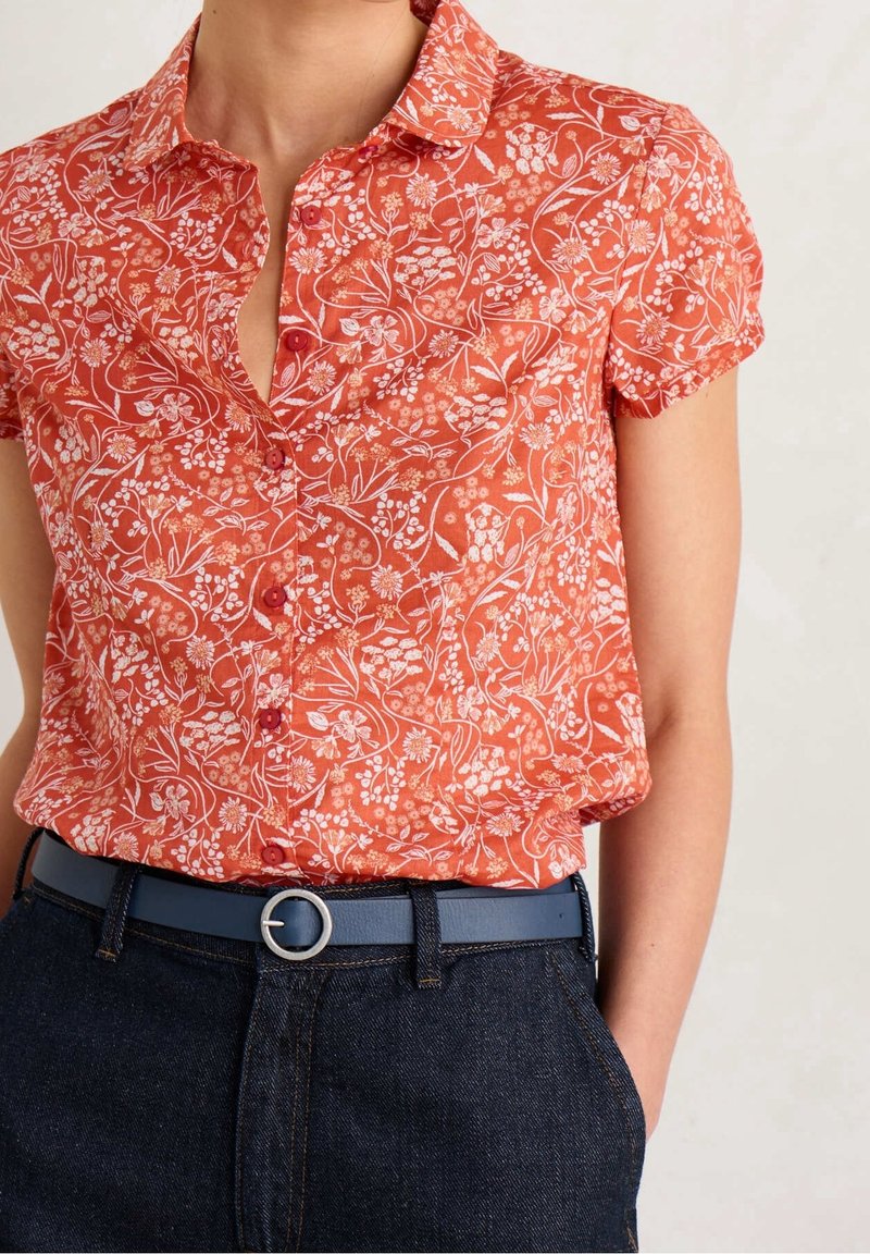 Camicia a maniche corte, con bottoni, di colore arancione con motivo floreale bianco. Abbinata a jeans scuri e una cintura blu con fibbia rotonda.