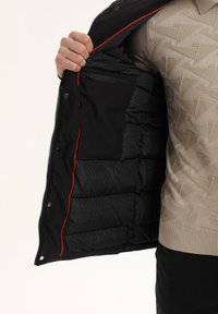 Chaqueta de plumas negra con cierre de cremallera, que presenta un forro interior rojo brillante y un bolsillo interior, mostrando una textura acolchada.
