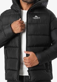 Homme portant une veste matelassée noire avec capuche, en train de la dézipper pour révéler un pull en tricot blanc en dessous.