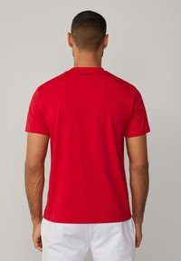 Camiseta roja de manga corta con cuello redondo. Presenta una textura suave y un logo negro en la parte superior de la espalda. Sin patrones ni acentos visibles.