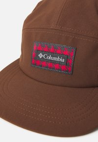 Hnědá látková čepice s prošitým okrajem, který nese obdélníkovou nášivku loga Columbia orámovanou červeným a černým vzorem houndstooth.