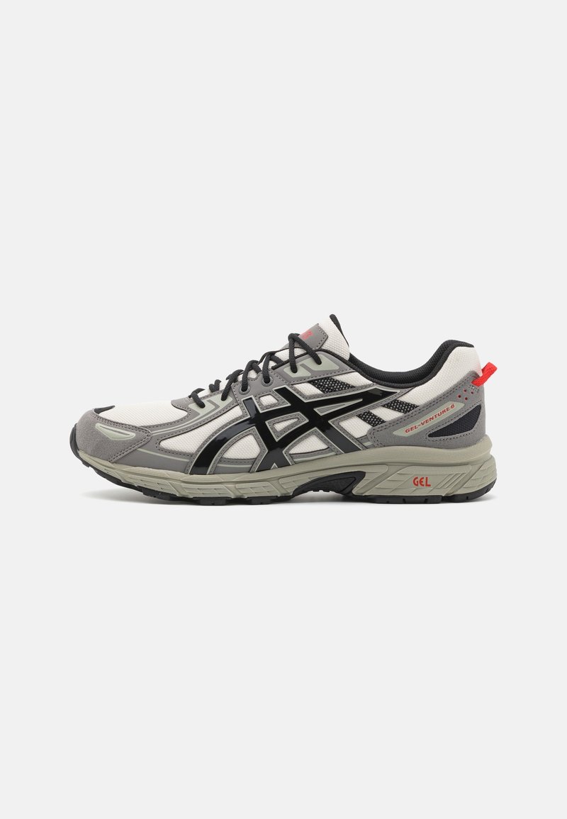 Chaussure de sport grise et blanche avec des accents noirs, lacets noirs et languette rouge, vue de côté sur fond blanc.
