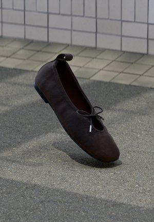 Ballerine en daim marron foncé avec semelle noire et petit nœud, posée sur un sol pavé près d’un mur carrelé.