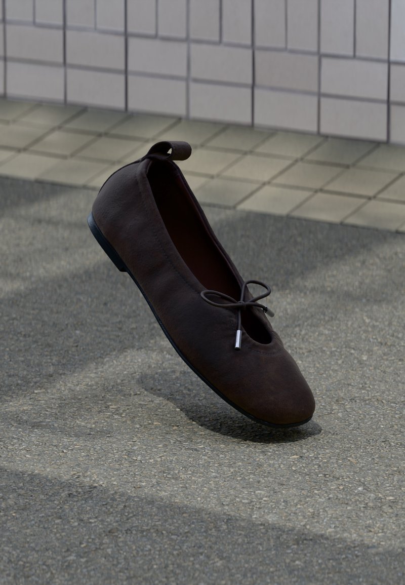 Σκούρο καφέ μπαλαρίνα από σουέτ με μαύρη σόλα και μικρό φιόγκο, που βρίσκεται σε πλακόστρωτο έδαφος κοντά σε πλακιδωτό τοίχο.