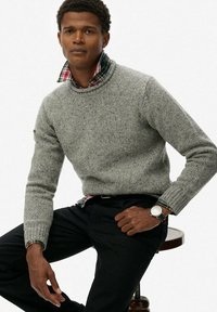 Suéter de punto gris con cuello redondo, con una textura moteada. Debajo, un cuello de camisa a cuadros en rojo, verde y azul. Pantalones negros y reloj en la muñeca.