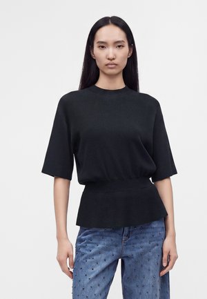 Femme asiatique aux longs cheveux noirs portant un haut noir ample à manches courtes et un jean bleu avec de petites perles noires, sur un fond blanc uni.