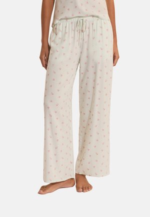 Mujer vistiendo pantalones de pijama beige claro con un pequeño estampado floral rosa y cintura con cordón, de pie descalza frente a un fondo blanco.