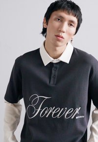 Camisa polo negra de manga corta con un collar blanco, con la palabra "Forever" en una fuente cursiva y "MISHY" en un texto más pequeño.