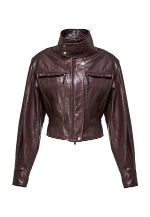 Žena u tamnoljubičastoj kožnoj jakni, crnoj suknji dužine do koljena, prozirnim čarapama i crnim čizmama do koljena s potpeticama, stoji na bijeloj pozadini.