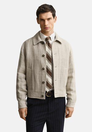 Man draagt een beige jasje met knopen over een wit shirt met een bruin gestreepte stropdas en donkere broek met krijtstrepen, staand tegen een witte achtergrond.