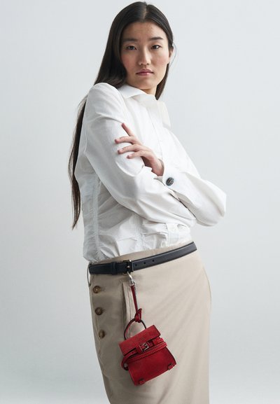 Κόκκινη σουέντ μίνι τσάντα με λαβή και λουράκι, διακοσμημένη με κούμπωμα. Συνδυασμένο με λευκό πουκάμισο και μπεζ φούστα με κούμπια.