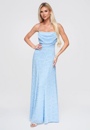 Blonde Frau in einem hellblauen, ärmellosen, bodenlangen Kleid mit dezentem Muster, posierend mit einer Hand in der Hüfte und der anderen auf dem Oberschenkel.
