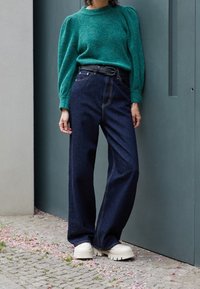 Tyrkis stickad tröja med puffärmar, kombinerad med högmidjade mörk denimjeans och klumpiga beige plattformsskor. Minimalistisk design.