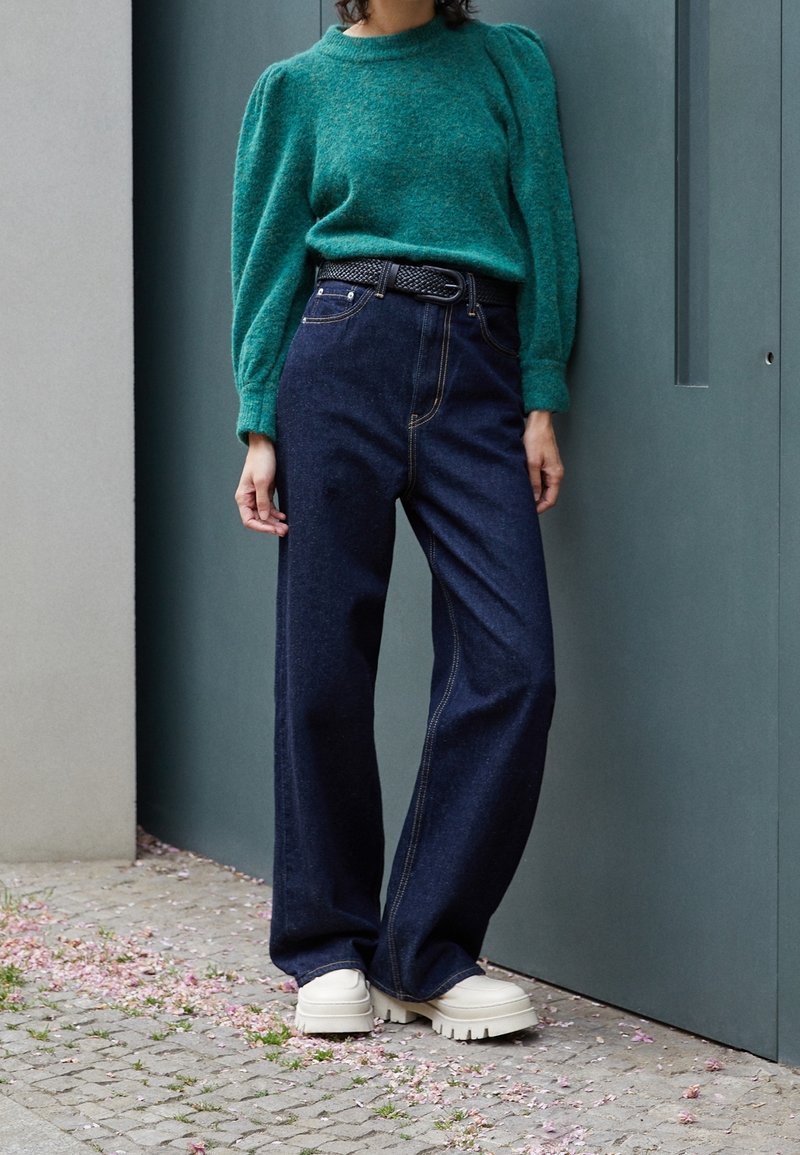 Tyrkis stickad tröja med puffärmar, kombinerad med högmidjade mörk denimjeans och klumpiga beige plattformsskor. Minimalistisk design.