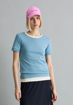 Młoda kobieta z krótkimi blond włosami nosi różową czapkę, jasnoniebieski t-shirt nałożony na biały top oraz czarną spódnicę, stoi na tle szarego tła.