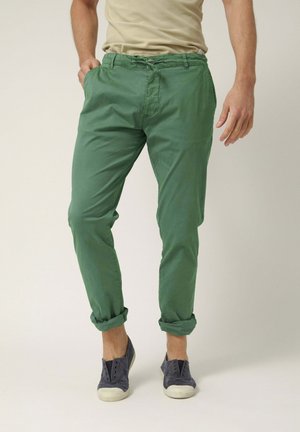 Hombre con pantalones verdes remangados, camisa beige y zapatos sin cordones grises, de pie con una mano en el bolsillo contra un fondo liso.
