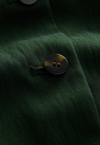Zielona tkanina o gładkiej fakturze i drobnych prążkach, z wyraźnym ciemnym guzikiem z dwoma otworami przyszytym nitką.