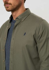 Camicia verde oliva con bottoni, dotata di colletti, con bottoni marroni e un piccolo logo blu navy. Indossata sopra un maglione a girocollo blu navy con texture.