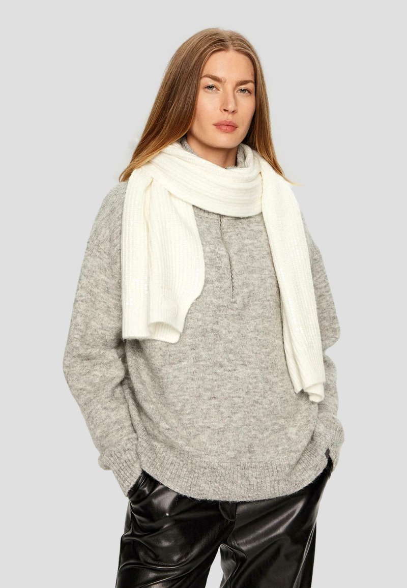 Maglione grigio a maglia con collo alto, abbinato a una sciarpa bianca a coste. Il modello indossa pantaloni neri in ecopelle lucida.