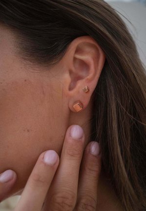Close-up of ear with two gold square stud earrings, one larger on earlobe and one smaller above, with fingers gently touching the neck.