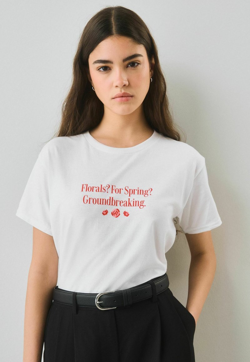 Jeune femme aux longs cheveux foncés portant un t-shirt blanc avec un texte rouge « Florals? For Spring? Groundbreaking. » et un pantalon noir avec une ceinture noire.