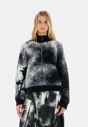 Maglione zip-up fluffy bianco e nero con colletto alto, caratterizzato da un motivo astratto e polsini a coste. Indossato sopra una gonna con fantasia.