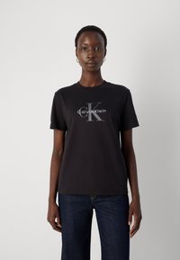 T-shirt en coton noir avec un logo Calvin Klein gris, col rond et manches courtes. Porté avec un jean foncé sur un fond clair.
