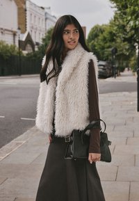 Kunstpelz-Weste in Weiß, braunes Langarmshirt und schwarze Handtasche. Model steht auf dem Bürgersteig, im Hintergrund Bäume und Gebäude.