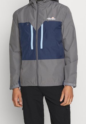 Homme portant une veste grise et bleu marine avec des cordons bleu clair et une fermeture éclair à l'avant, associée à un pantalon noir.