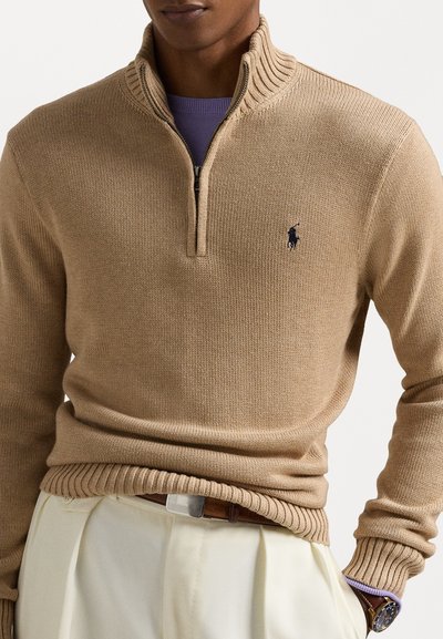 Beige stickad ziptröja med ribbad krage och nederkant, prydd med en mörk logotyp på vänster bröst och lagd över en lila skjorta.