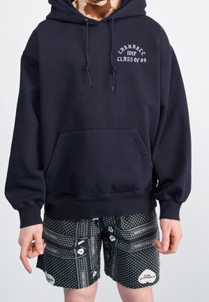 Person wearing a black Carhartt hoodie with "MIP Class of 89" text and black patterned shorts with white geometric shapes.