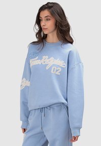 Sweatshirt oversize bleu clair à manches longues, avec un texte brodé blanc "True Religion 02" et un emblème circulaire sur la poitrine.