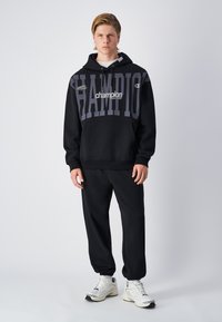 Felpa nera con grande scritta grigia "CHAMPION", cappuccio con cordini, tasca frontale. Abbinata a pantaloni della tuta neri e sneakers bianche. Abbigliamento sportivo casual.