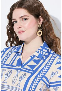 Camicia a maniche corte con base color crema e motivi di pesci e foglie blu. Presenta un colletto e una texture morbida. Orecchini dorati e intrecciati aggiungono dettagli.