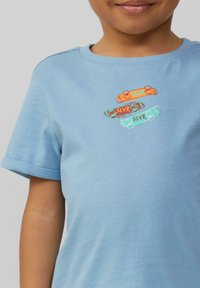 Lichtblauwe katoenen T-shirt met drie kleurrijke skateboardgraphics in oranje, bruin en teal, met de tekst "SLVR". Omgeslagen mouwen.