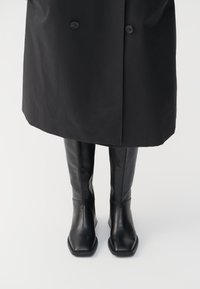 Botas de cuero negras hasta la rodilla con punta cuadrada, textura suave y diseño minimalista, combinadas con un abrigo largo negro.