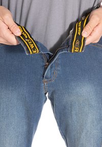 Jean en denim bleu avec des bretelles jaunes et noires, fermeture éclair exposée et coutures orange le long des coutures, maintenu à la taille.