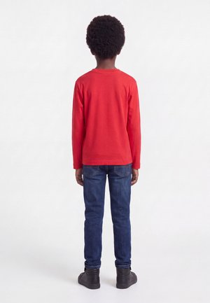 Niño con pelo rizado vestido con una camiseta roja de manga larga, vaqueros azules y zapatos negros, de pie mirando hacia atrás sobre un fondo blanco.