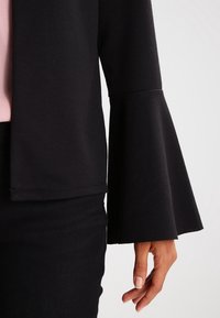 Svart topp med klockärmar i mjukt tyg, med en slät textur och en figurnära passform. Lagerdesign med subtila detaljer längs fållen.