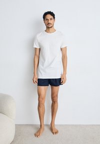 T-shirt en coton blanc à manches courtes associé à un short bleu foncé ; se tenant sur un tapis clair devant un mur uni, pieds nus.