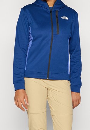 Osoba nosząca niebieską bluzę z kapturem na zamek błyskawiczny z kieszenią na piersi oraz beżowe, rozpinane spodnie trekkingowe, stojąca na jednolitym tle.