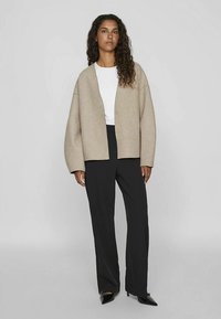 Beżowy, oversizeowy kardigan z zaokrąglonymi ramionami, zapinany z przodu na guziki, noszony na białej koszulce i czarnych szerokich spodniach, uzupełniony czarnymi szpilkami.