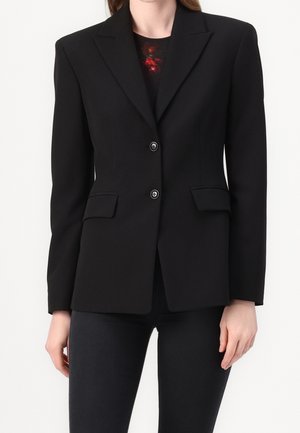Vrouw met een strakke zwarte blazer met inkeping in de revers, twee knopen, klepzakken en een zwarte broek, zichtbaar zijn bovenlichaam en armen.