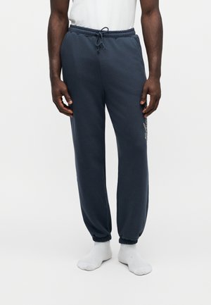 Mann mit marineblauen Jogginghosen mit elastischen Bündchen und Kordelzug in der Taille, kombiniert mit weißen Socken vor weißem Hintergrund.
